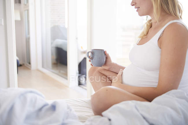 Cropped shot of pregnant young woman sitting on bed with coffee — Stock Photo