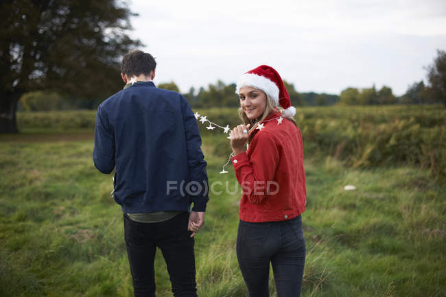 Visão traseira do jovem casal em santa chapéu e guirlanda leve passeando no campo — Fotografia de Stock