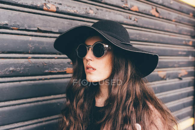 Portrait of young woman standing beside shutter, wearing floppy hat and sunglasses — Stock Photo