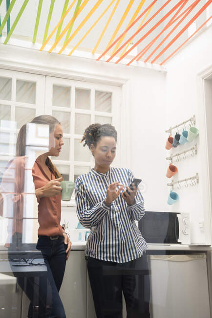 Two businesswomen looking at smartphone while taking a coffee break in office kitchen — Stock Photo