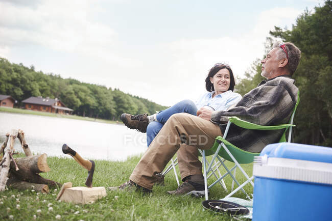 Casal maduro sentado em cadeiras de acampamento ao lado do lago — Fotografia de Stock