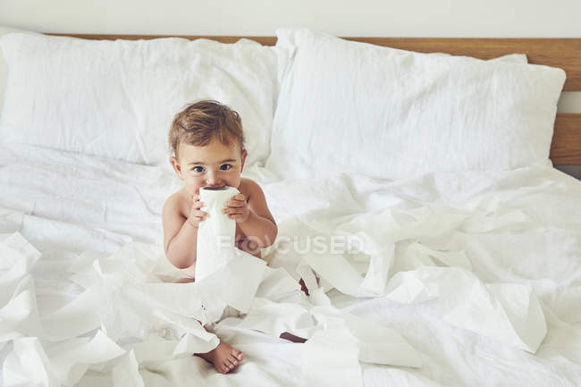 Тоддлер сидит на кровати, держа распутанный рулон унитаза — стоковое фото