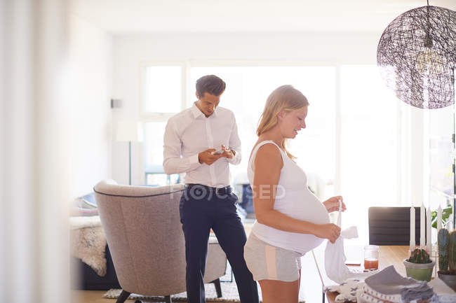Мужчина смотрит на смартфон и беременную девушку, складывающую белье в гостиной — стоковое фото