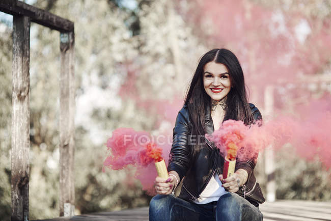 Young woman sitting on decking, holding smoke flares, smoke pouring from flares — Stock Photo