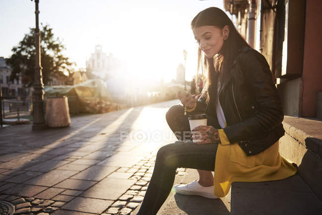 Молодая женщина на улице, держит чашку кофе и смартфон, татуировки на руках — стоковое фото