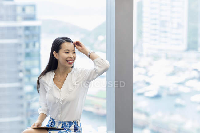 Mujer con tableta digital sentada en un alféizar mirando hacia otro lado sonriendo - foto de stock