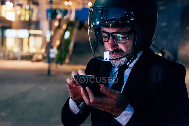 Mature businessman in motorcycle helmet using smartphone at night — Stock Photo