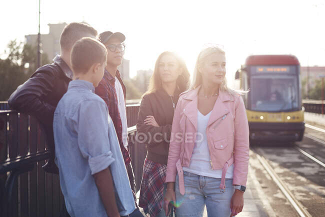 Cinco amigos adultos jóvenes esperando en la estación de tranvía de la ciudad iluminada por el sol - foto de stock