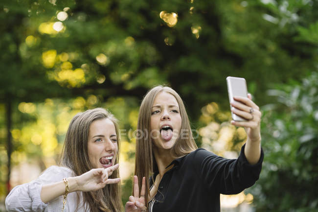 Two young female friends pulling faces for smartphone selfie in park — Stock Photo