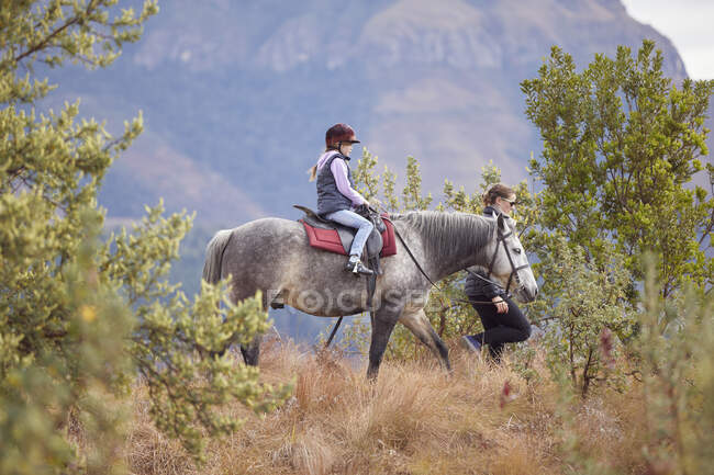Menina equitação cavalo em ambiente rural, mãe andando ao seu lado — Fotografia de Stock