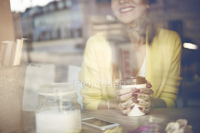 Mujer joven sentada en la cafetería, sosteniendo la taza de café, tatuajes a mano, vista a través de la ventana de la cafetería, sección central - foto de stock
