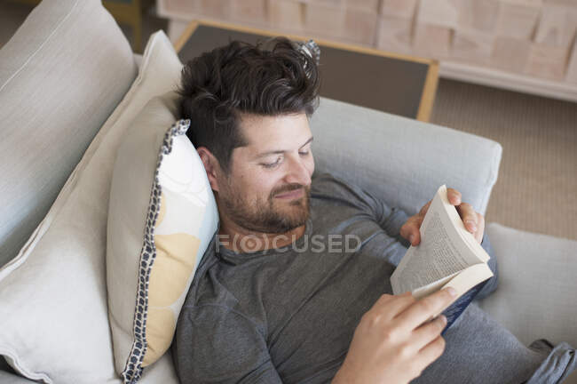 Середній дорослий чоловік розслабляється на дивані, читає книгу, підвищений вид — стокове фото