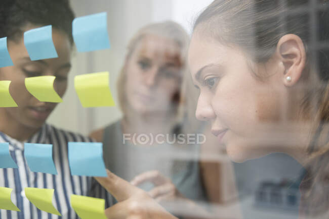 Three businesswomen behind glass wall pointing at adhesive notes — Stock Photo