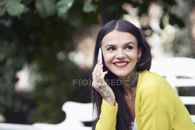 Jeune femme assise à l'extérieur, utilisant un smartphone, souriant, tatouages sur la main et le cou — Photo de stock