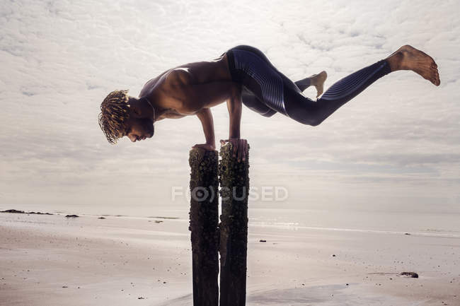 Молодой человек делает стойку на руках на деревянных столбах пляжа — стоковое фото