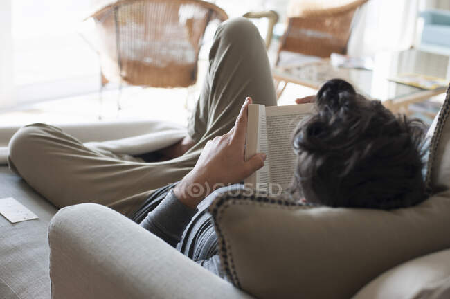 Середній дорослий чоловік розслабляється на дивані, читає книгу, вид ззаду — стокове фото