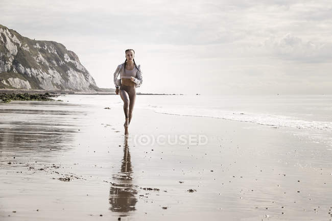Vista frontal de una joven corredora corriendo descalza por la playa - foto de stock