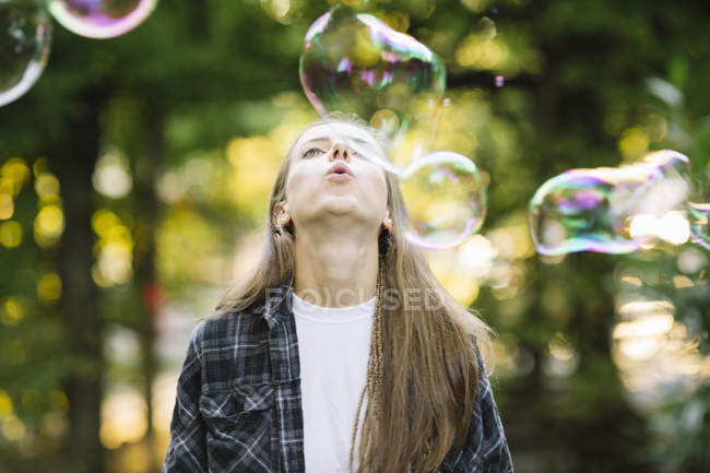 Mujer joven soplando burbuja flotante hacia arriba en el parque - foto de stock