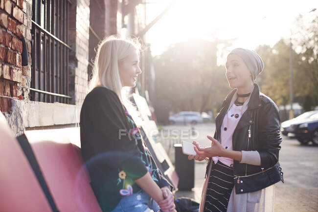 Dos amigas jóvenes charlando en la calle de la ciudad iluminada por el sol - foto de stock