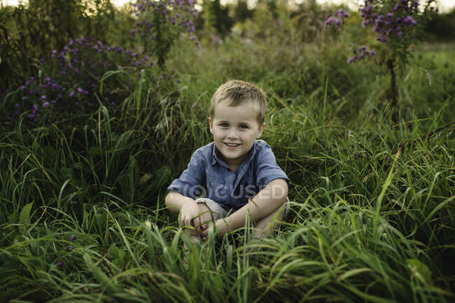 Porträt eines lächelnden Jungen, der im hohen Gras sitzt und in die Kamera blickt — Stockfoto