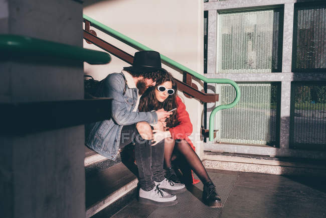Молодая пара сидит на ступеньках, смотрит на смартфон — стоковое фото