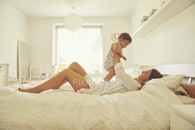 Мати лежить на ліжку, тримає малюка вгору, посміхається — стокове фото
