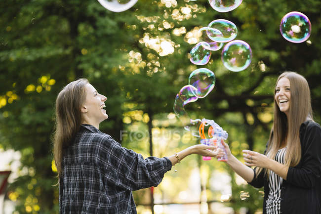 Two young female friends making floating bubbles in park — Stock Photo