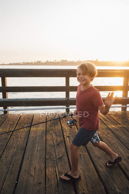 Niño en el muelle con caña de pescar saludando a la cámara sonriendo, Goleta, California, Estados Unidos, América del Norte - foto de stock