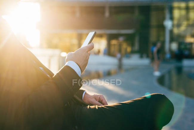 Maduro hombre de negocios sentado al aire libre, utilizando el teléfono inteligente, sección media - foto de stock
