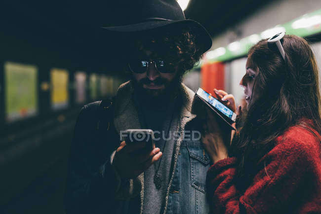 Pareja joven sentada en la plataforma del metro, mirando los teléfonos inteligentes - foto de stock