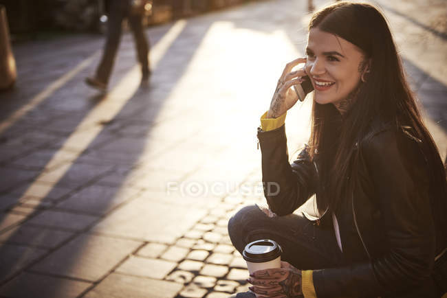 Mujer joven sentada al aire libre, sosteniendo la taza de café, usando un teléfono inteligente, tatuajes en las manos - foto de stock