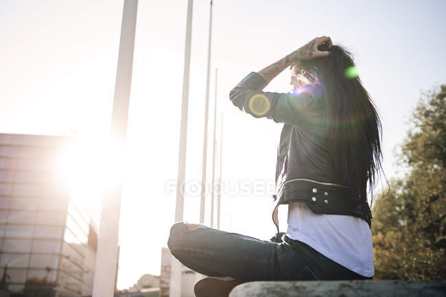 Mujer joven sentada con las piernas cruzadas en la pared, frente a la luz del sol, tatuajes en la mano - foto de stock
