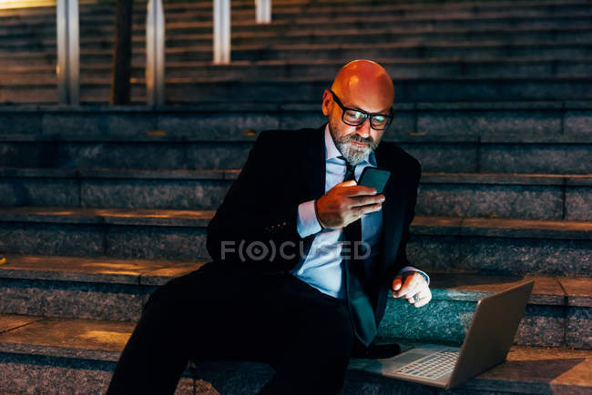 Mature businessman sitting on steps with smartphone and laptop — Stock Photo