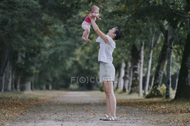 Woman holding up baby daughter in tree lined park — Stock Photo