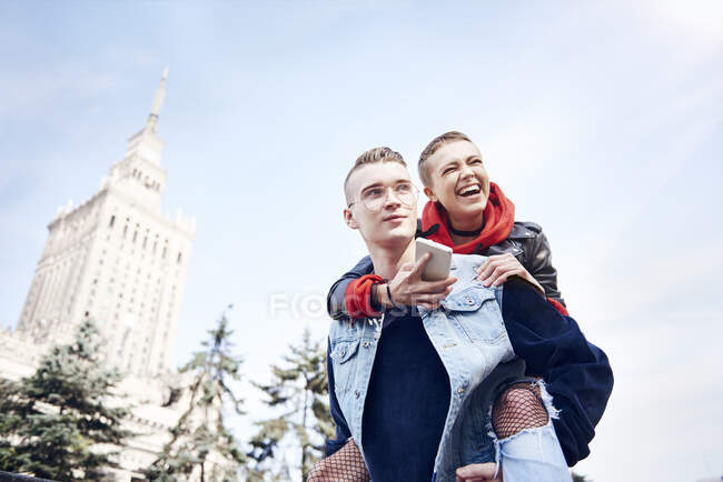 Joven hombre dando riéndose novia un cerdito de nuevo en la ciudad - foto de stock