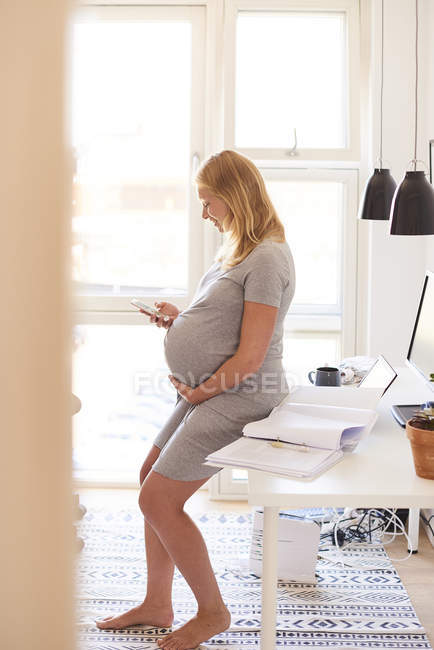 Pregnant woman leaning against desk and looking at smartphone — Stock Photo