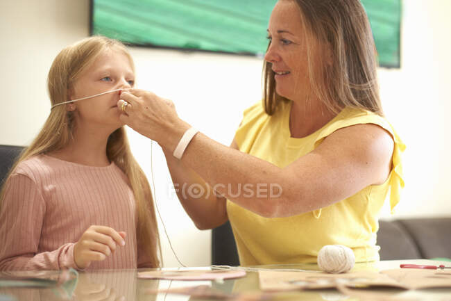Madre e hija sentadas a la mesa, madre midiendo el rostro de la hija con cuerda - foto de stock