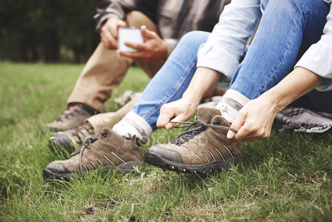 Pareja madura sentada en la hierba y la mujer atando botas a pie - foto de stock
