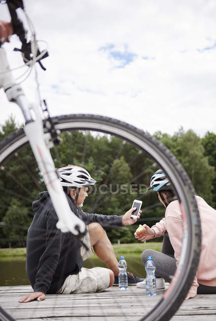 Mature couple in bicycle helmets looking at smartphone on pier — Stock Photo