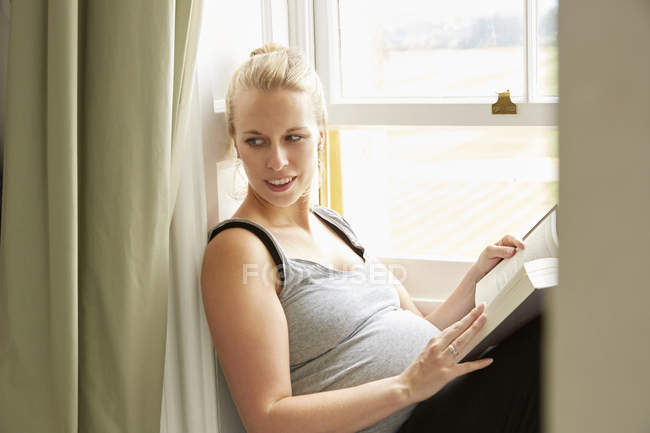 Donna incinta con libro seduta vicino alla finestra — Foto stock