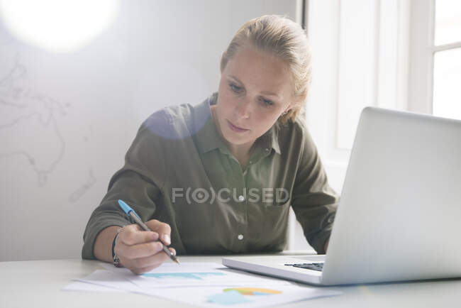 Jovem empresária fazendo anotações e usando laptop na mesa de escritório — Fotografia de Stock