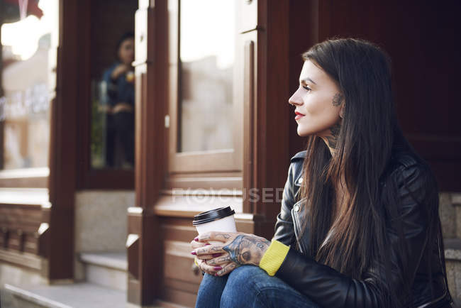 Jeune femme assise à l'extérieur, tenant une tasse de café, tatouages sur les mains — Photo de stock