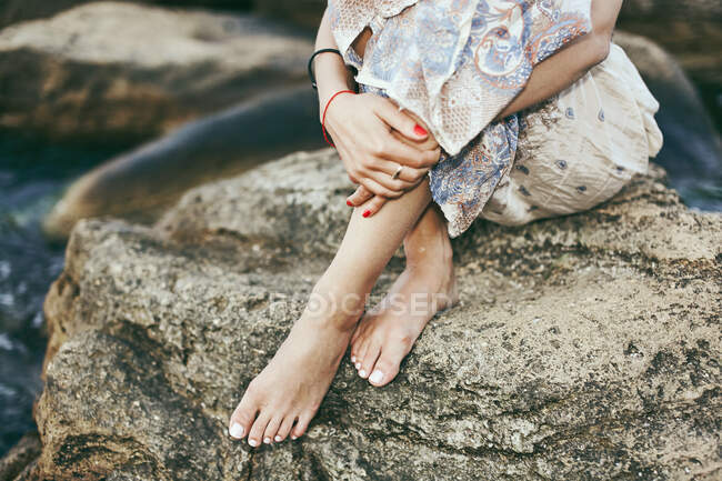 Vista de la cintura hacia abajo de la joven sentada descalza en la roca costera, Odessa, Ucrania - foto de stock