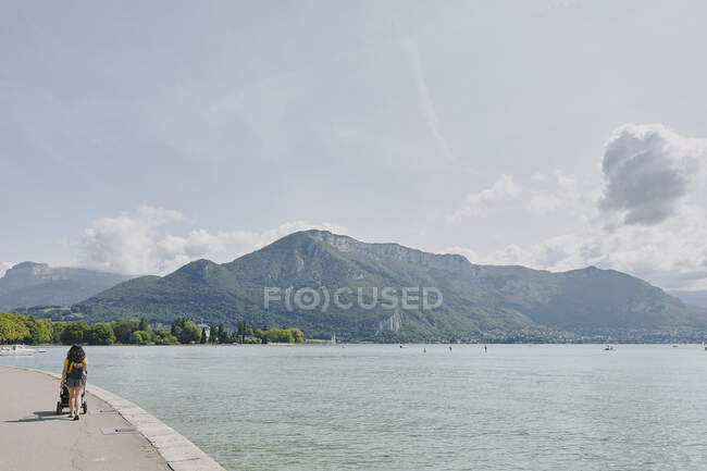 Rear view of woman strolling with pushchair waterfront at Lake Annecy, Annecy, Auvergne-Rhone-Alpes, France — Stock Photo