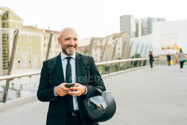 Homem de negócios maduro na ponte segurando smartphone e capacete de motocicleta — Fotografia de Stock