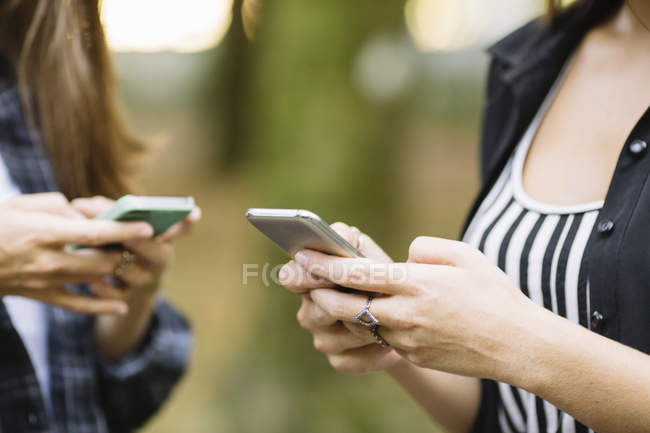 Tiro cortado de duas jovens mulheres usando tela sensível ao toque do smartphone no parque — Fotografia de Stock