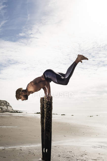 Young man doing handstand with raised legs on wooden beach posts — Stock Photo