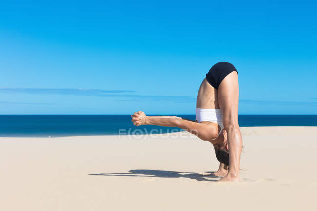 Vista lateral da mulher na praia se curvando para a frente na posição de ioga — Fotografia de Stock