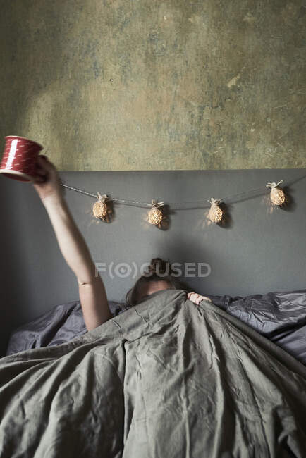 Mulher na cama, escondido sob coberturas, segurando caneca no ar — Fotografia de Stock