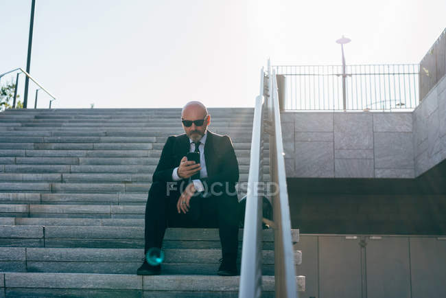 Mature businessman sitting on steps and using smartphone — Stock Photo